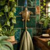 Brass sea turtle wall hook mounted on a green tiled wall holding a towel among plants, handmade nautical coastal coat hook.