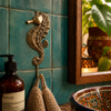 Solid brass seahorse wall hook mounted on teal glazed bathroom tiles beside a wooden mirror with linen towel and Aesop products — coastal beach house bathroom hook handmade in Bali