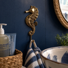 Solid brass seahorse wall hook mounted on a navy blue bathroom wall beside a round mirror with striped towel hanging and soap dispenser — nautical brass coat hook beach house decor handmade in Bali