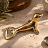 Gold brass seal bottle opener resting on a sandy stone surface beside scattered bottle caps and green plants. A nautical bar decor piece and beer bottle opener for a home bar or man cave, handmade in Bali.