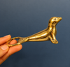 Brass seal bottle opener held in a hand against a dark blue background, showing its compact size and polished gold finish. A pocket-sized bartender gift and seal bottle opener, handmade in Bali.