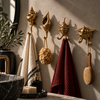 Set of 4 brass seashell wall hooks mounted in a row on a bathroom wall, holding cream and burgundy towels and a natural loofah beside a mirror and green plants. Handmade cast brass shell towel hooks for a coastal, nautical or beach house bathroom, made in Ubud, Bali.
