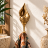 Brass shell hook shaped like an open spiral conch seashell with Balinese Batik-style carving, mounted on a cream bathroom wall and holding a patterned towel beside a stone basin and dried flowers. Handmade cast brass shell towel hook for a coastal or nautical bathroom, hand-finished in Ubud, Bali.