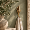 Brass spiral seashell towel hook with Balinese carving holding a cream towel on a sage green wall, beside a stone vase with olive branches. Handmade cast brass coastal bathroom hook, hand-finished by artisans in Ubud, Bali.