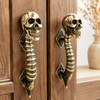 Solid brass skull spine door handle pair installed on warm wood cabinet showing detailed vertebrae skull face and diamond mounting plate — gothic double door pull handmade in Ubud Bali