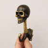 Hand holding solid brass skull wall hook in dark antique finish from front showing full skeleton figure skull ribcage spine and curved hook for scale — skeleton hook handmade in Ubud Bali