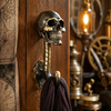 Solid brass skull wall hook mounted on a dark gothic steampunk wall with purple scarf hanging beside vintage lantern and clockwork decor — gothic skull coat hook handmade in Ubud Bali