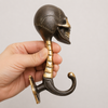 Hand holding solid brass skull wall hook in dark antique finish at an angle showing skull face spine and curved hook from below — gothic skull coat hook handmade in Ubud Bali