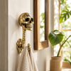 Small solid brass skull wall hook in polished gold finish mounted in a bright bathroom with white towel hanging beside a mirror and monstera plant — gothic skull towel hook clearance handmade in Ubud Bali