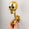 Hand holding small solid brass skull wall hook in polished gold finish showing full skeleton figure skull ribcage spine and curved hook for scale — gothic skull coat hook handmade in Ubud Bali