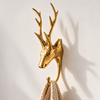 Slim brass stag wall hook with tall upright antlers holding a beige knit towel on a cream wall with soft shadows. One of the three designs in the brass deer wall hooks set of 3, a handmade towel hook and coat hook cast in Bali.