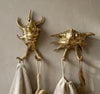 Two brass spiky conch shell double hooks, one upright and one sideways, mounted on a warm beige wall holding a cream towel and a drawstring bag. Handmade cast brass seashell towel and coat hooks with two hanging points each, from the brass seashell wall hook set of 4.