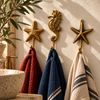 Brass starfish and seahorse wall hook set of 3 mounted in a row on a cream bathroom wall, holding rust, navy and striped towels beside a stone basin and green plants. Handmade cast brass sea star and seahorse towel hooks for a coastal, nautical or beach house bathroom, made in Ubud, Bali.