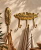 Gold brass feather wall hook and 3-hook feather coat rack on a sunlit cream wall, holding a woven straw bag and a striped cotton towel beside green plants. Handmade cast brass boho entryway hooks in a polished gold finish, hand-cast and finished by artisans in Ubud, Bali.