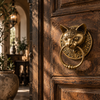 Gothic door knocker in antique brass mounted on a carved wooden door in a warm candlelit hallway, shaped like a cat head with an ornate ring. A handmade brass cat head door knocker and dark academia decor piece, a unique cat lover gift from Ubud, Bali.