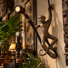 Hanging monkey sconce in antique brass on a warm wall in a Bali-style interior with carved wooden panels, a tribal mask and tropical plants, its spotlight glowing. A handmade brass monkey wall lamp bringing jungle decor and statement lighting to a living room or bar.