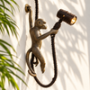 Hanging monkey wall lamp in antique brass facing the opposite direction on a cream wall with palm leaf shadows, the monkey gripping a styling vine and holding the spotlight. A handmade brass plug-in sconce and statement tropical light from Ubud, Bali.