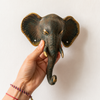 Hand holding patina brass elephant wall hook showing size scale, rich verdigris patina finish, golden tusk detail and warm gold ear accents against clean white background. Handcrafted decorative coat hook and towel hook from set of 4 for boho home decor, wabi sabi decor, yoga room decor and eclectic gallery wall decor. Unique boho housewarming gift set of 4 with no tariffs for US buyers
