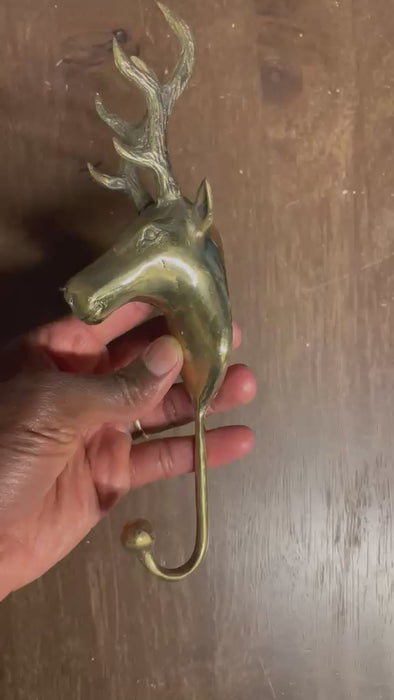 Video of the rustic brass reindeer wall hook held in a hand against a wooden door, turned to show the stag's head, branching antlers and curved hook. Handmade brass deer hook from Ubud, Bali.