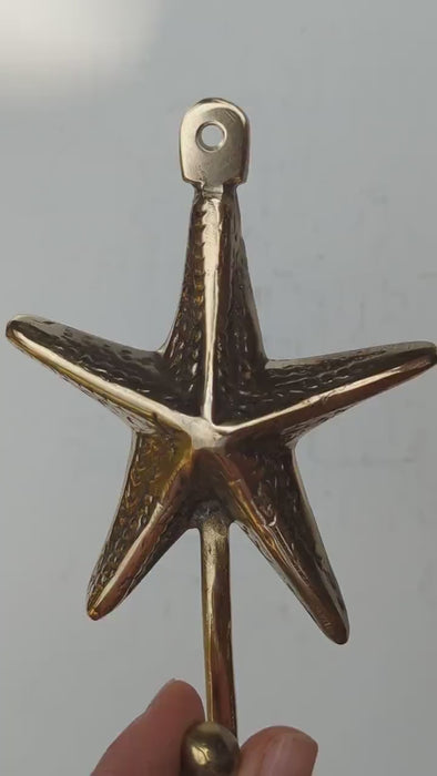 Video close-up of the brass textured starfish wall hook held in a hand, showing the detailed sea star arms, the top screw loop and the ball-tipped hook. Handmade cast brass coastal hook from Ubud, Bali.