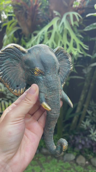 Hand holding handcrafted patina brass elephant wall hook outdoors in tropical garden setting showing rich verdigris patina finish and golden tusk and ear detail highlighting Bali artisan craftsmanship. Decorative coat hook and towel hook from set of 4 for boho home decor, wabi sabi decor, yoga room decor and meditation space decor. Unique boho housewarming gift and eclectic gallery wall hook set