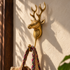 Rustic brass reindeer wall hook in polished gold, mounted on a sunlit cream wall beside a dark wooden beam and holding a woven bag. A handmade deer wall hook with tall branching antlers that works as an antler coat hook or rustic wall hook, hand-cast in Bali.
