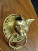 Angled view of the brass cat head on a wooden surface, showing the profile of the cat's face, its pointed ears and the brass ring resting on the ornate plate. A handmade drawer pull, towel ring and door knocker from Ubud, Bali.