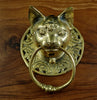 Polished gold brass cat head towel ring on a dark wooden surface, the warm brass catching the light on the cat's face and scrollwork plate. A handmade brass towel ring and gothic door knocker for dark academia homes, made in Ubud, Bali.